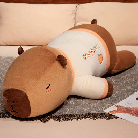 Plush capybara toy resembling a sloth on a bed with visible brand name.