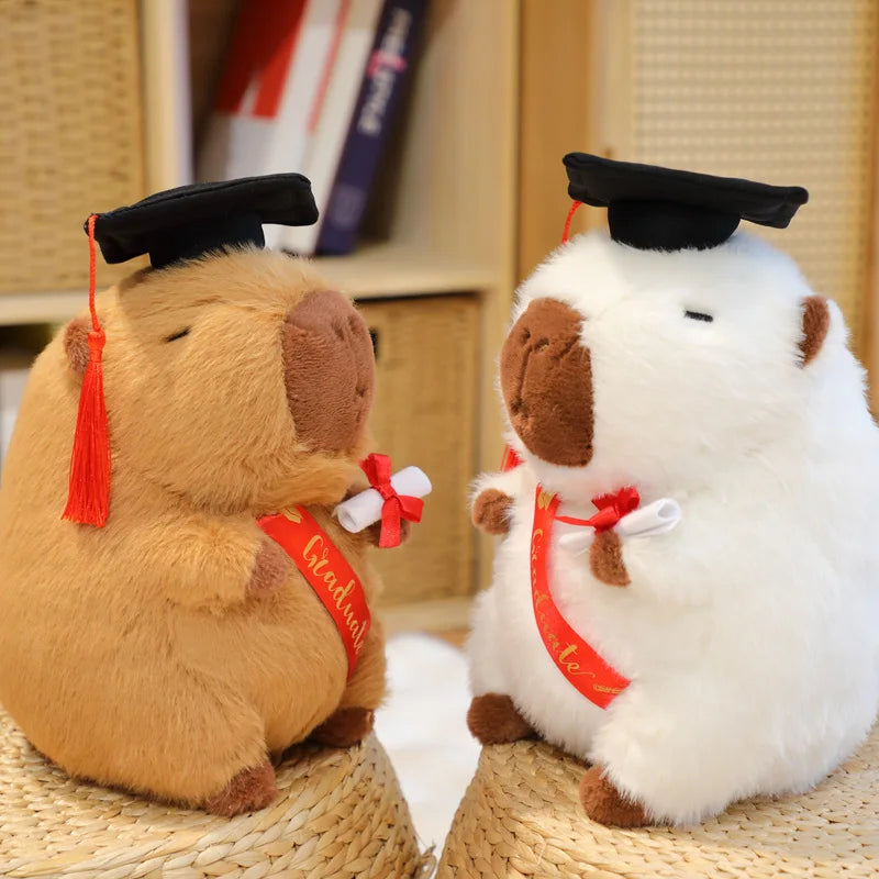 Two plush capybara toys, one brown and one white, wearing graduation caps and gowns on a woven surface.