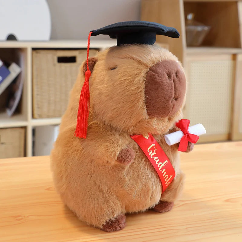 Plush toy resembling a capybara wearing a graduation cap and diploma on a wooden surface.