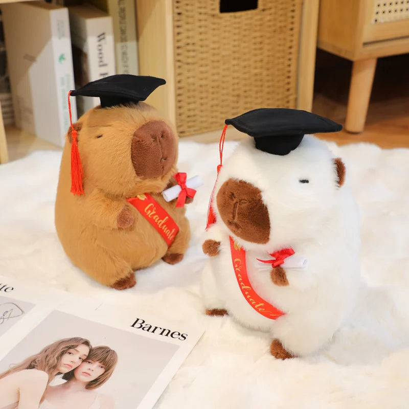 Two plush toys resembling capybaras in graduation caps and gowns on a surface with a magazine underneath.